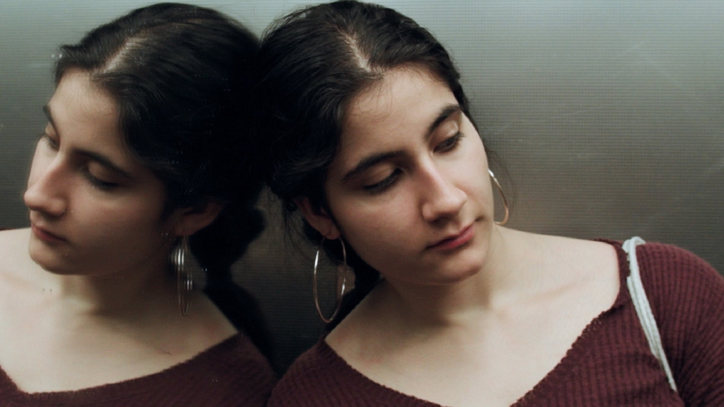 "Köy": Eine junge Frau mit schwarzen, zusammengebundenen Haaren, großen Ohrringen und dunkelrotem Pullover lehnt an der verspiegelten Wand einer Aufzugskabine, so dass man sie doppelt sieht.