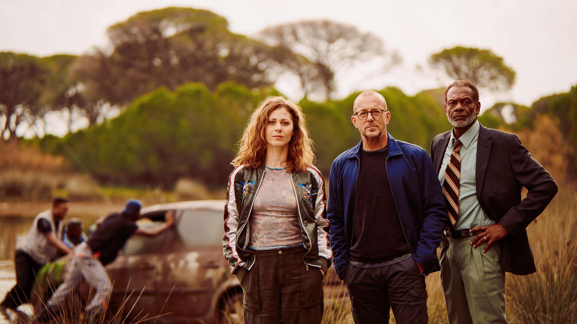 "Tod in Mombasa": Moritz Wagner (Heino Ferch), Caroline Jakobs (Katharina Schlothauer) und Kommissar Lomboto (Errol Trotman Harewood) stehen nebeneinander und schauen ernst in die selber Richtung. Hinter ihnen befindet sich ein See, aus welchem drei Männer gerade ein dreckiges Auto schieben. Hinter dem See stehen Pinien und andere Bäume.