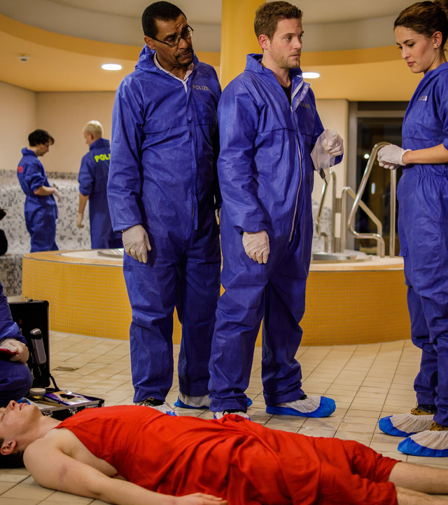 "SOKO München - Wann ist ein Mann ein Mann?": Am Tatort in einem Schwimmbad liegt eine Leiche auf dem Boden. Dr. Weissenböck (Florian Odendahl) schaut sich den Kopf der Leiche näher an. Franz Ainfachnur (Christofer v. Beau), Dominik Morgenstern (Joscha Kiefer) und Toni Bischoff (Amanda da Gloria) stehen daneben und beraten sich. Im Hintergrund sind Kollegen von der Spurensicherung (Komparsen) zu sehen.