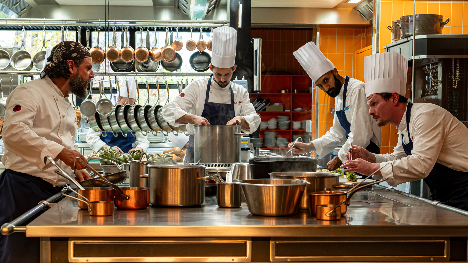 "München Mord - Im Zweifel für den Zweifel": Maximilian Dobek (Manuel Cortez) steht gemeinsam mit Oliver Gerg (Shenja Lacher) und zwei Beiköchen (Komparsen) in einer professionellen Küche. Alle tragen weiße Kochjacken und klassische Kochmützen. Vor ihnen erstreckt sich ein großer Arbeitstisch, auf dem zahlreiche Töpfe und Pfannen dampfen und brutzeln – jeder ist konzentriert bei der Sache. Im Hintergrund hängen Küchenutensilien ordentlich an Haken.