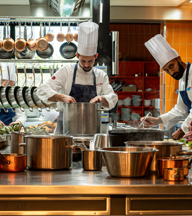 "München Mord - Im Zweifel für den Zweifel": Maximilian Dobek (Manuel Cortez) steht gemeinsam mit Oliver Gerg (Shenja Lacher) und zwei Beiköchen (Komparsen) in einer professionellen Küche. Alle tragen weiße Kochjacken und klassische Kochmützen. Vor ihnen erstreckt sich ein großer Arbeitstisch, auf dem zahlreiche Töpfe und Pfannen dampfen und brutzeln – jeder ist konzentriert bei der Sache. Im Hintergrund hängen Küchenutensilien ordentlich an Haken.
