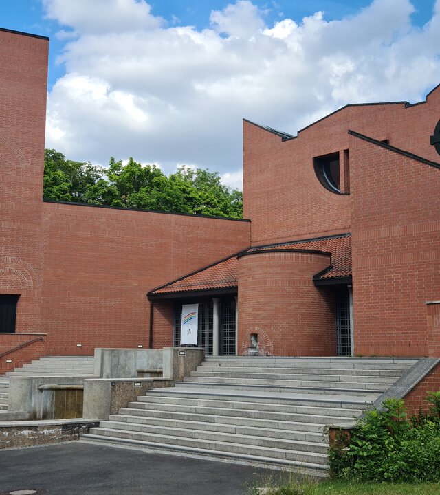 "Evangelischer Gottesdienst - Himmelhoch und tief verwurzelt": Außenansicht des Klosters Schwanberg in Unterfranken: Bei dem modernen, architektonisch interessant gestalteten Gebäude aus roten Backstein führt eine breite graue Treppe zum Eingang, der Turm ist etwas nach vorne versetzt.