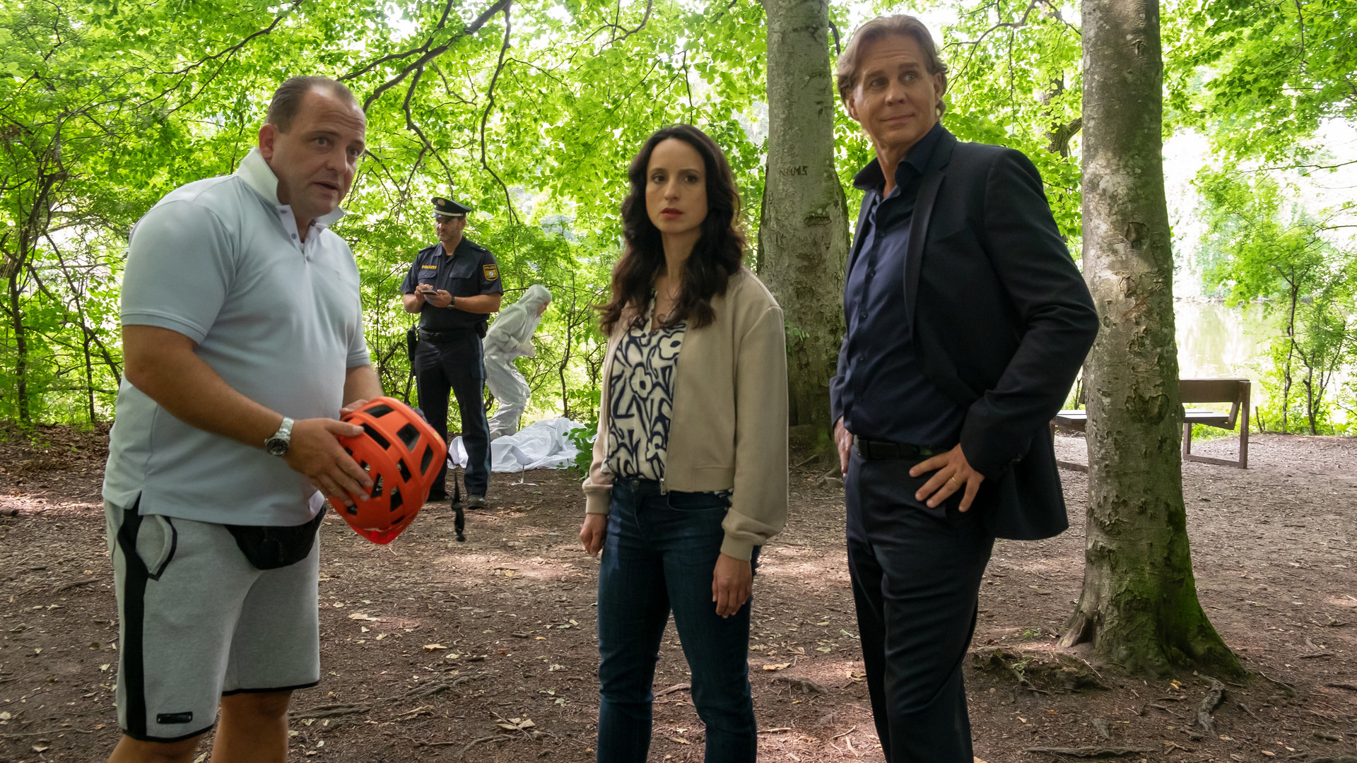 "Der Alte - Abstiegsangst": Simon Rothe (Urs Rechn) steht mit einem Fahrradhelm in der Hand im Wald. Annabell Lorenz (Stephanie Stumph) und Caspar Bergmann (Thomas Heinze) stehen daneben. Alle drei schauen in die Ferne. Im Hintergrund steht ein Polizist bei der Spurensicherung.