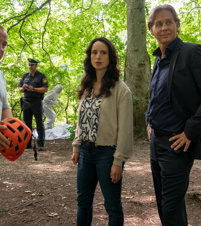 "Der Alte - Abstiegsangst": Simon Rothe (Urs Rechn) steht mit einem Fahrradhelm in der Hand im Wald. Annabell Lorenz (Stephanie Stumph) und Caspar Bergmann (Thomas Heinze) stehen daneben. Alle drei schauen in die Ferne. Im Hintergrund steht ein Polizist bei der Spurensicherung.