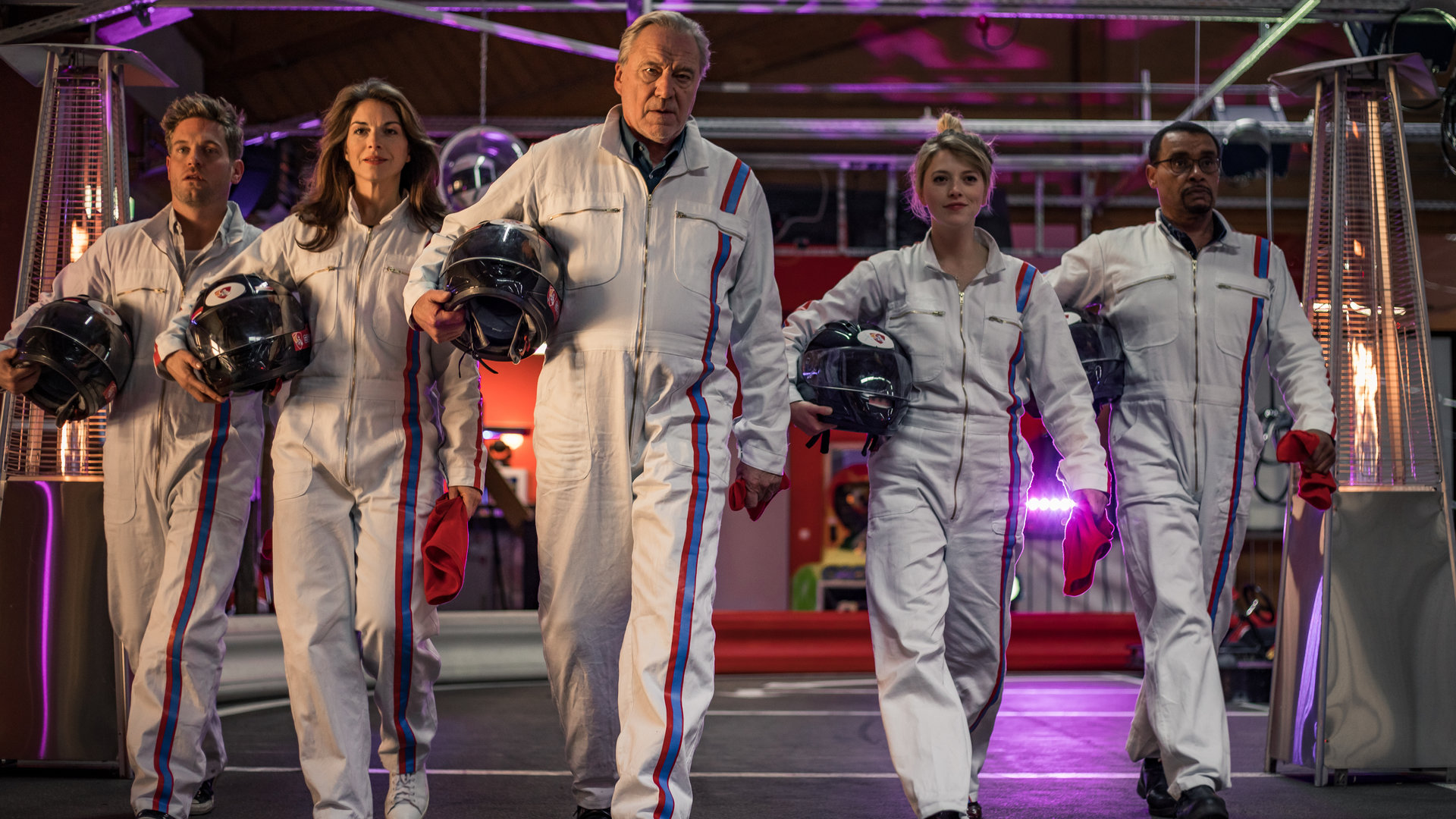 "SOKO München - Tod in Bestzeit": Katharina Hahn (Bianca Hein), Franz Ainfachnur (Christofer v. Beau), Arthur Bauer (Gerd Silberbauer), Dominik Morgenstern (Joscha Kiefer) und Resi Schwaiger (Mersiha Husagic) laufen nebeneinander über die Gokartbahn. Sie tragen einheitlich weiße Ganzkörperanzüge mit einem blauen und einem roten Streifen an der Seite. Unter ihren rechten Armen halten sie einen Schutzhelm. In der linken Hand trägt jeder ein rotes Stück Stoff.