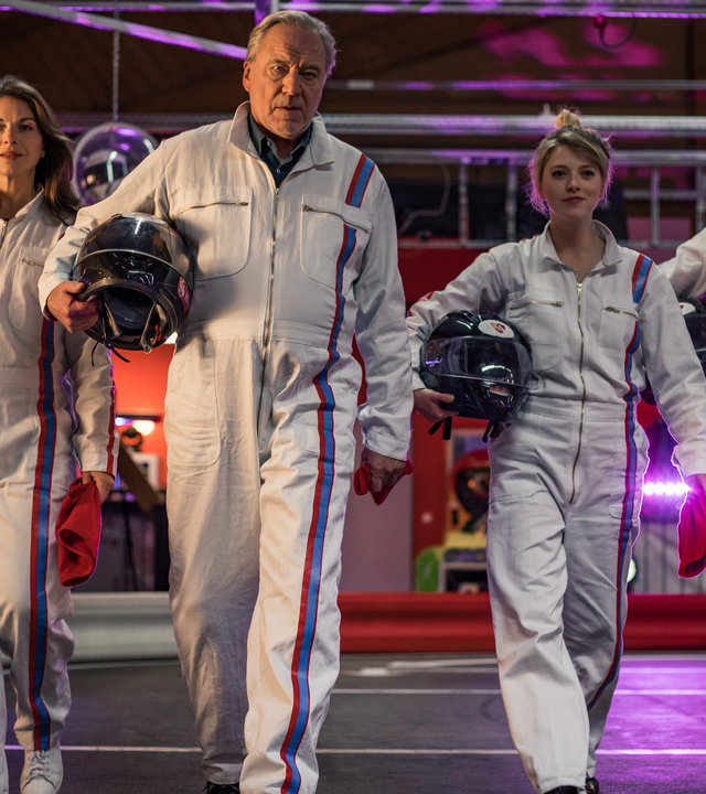 "SOKO München - Tod in Bestzeit": Katharina Hahn (Bianca Hein), Franz Ainfachnur (Christofer v. Beau), Arthur Bauer (Gerd Silberbauer), Dominik Morgenstern (Joscha Kiefer) und Resi Schwaiger (Mersiha Husagic) laufen nebeneinander über die Gokartbahn. Sie tragen einheitlich weiße Ganzkörperanzüge mit einem blauen und einem roten Streifen an der Seite. Unter ihren rechten Armen halten sie einen Schutzhelm. In der linken Hand trägt jeder ein rotes Stück Stoff.