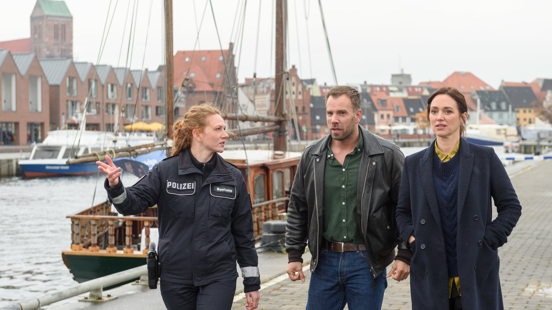 "SOKO Wismar – Junggesellenabschied": Paula Moorkamp (Stelle Hinrichs) führt ihre Kollegen Karoline Joost (Nike Fuhrmann) und Lars Pöhlmann (Dominic Boeer) an der Kaimauer entlang. Sie deutet außerhalb des Bildes zur Leiche. Im Hintergrund sind mehrere Boote im Hafenbecken, die St. Nikolai-Kirche und die Hafenfassade zu sehen.
