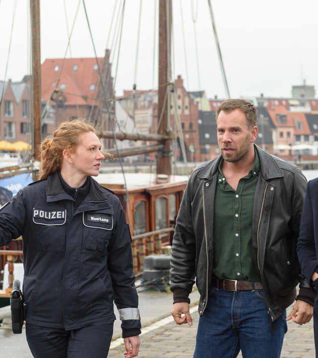 "SOKO Wismar – Junggesellenabschied": Paula Moorkamp (Stelle Hinrichs) führt ihre Kollegen Karoline Joost (Nike Fuhrmann) und Lars Pöhlmann (Dominic Boeer) an der Kaimauer entlang. Sie deutet außerhalb des Bildes zur Leiche. Im Hintergrund sind mehrere Boote im Hafenbecken, die St. Nikolai-Kirche und die Hafenfassade zu sehen.
