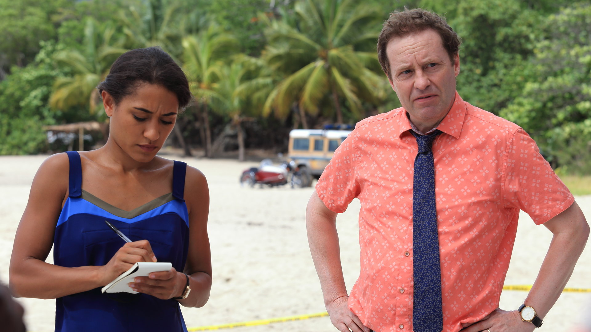 "Death in Paradise - Die Kunst des Schreibens": Florence Cassell (Joséphine Jobert) hat einen Block und Stift in den Händen DI Jack Mooney (Ardal O´Hanlon) steht daneben und stützt die Hände auf der Hüfte ab. Beide stehen am Strand.