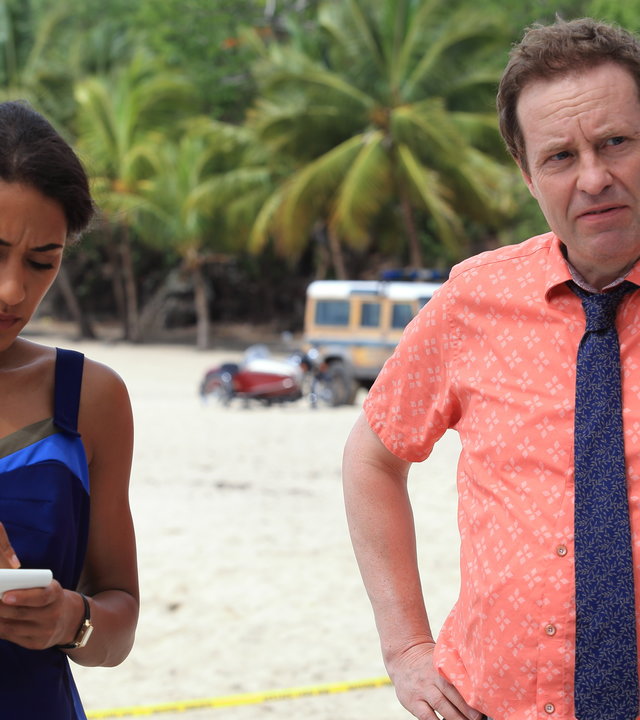 "Death in Paradise - Die Kunst des Schreibens": Florence Cassell (Joséphine Jobert) hat einen Block und Stift in den Händen DI Jack Mooney (Ardal O´Hanlon) steht daneben und stützt die Hände auf der Hüfte ab. Beide stehen am Strand.